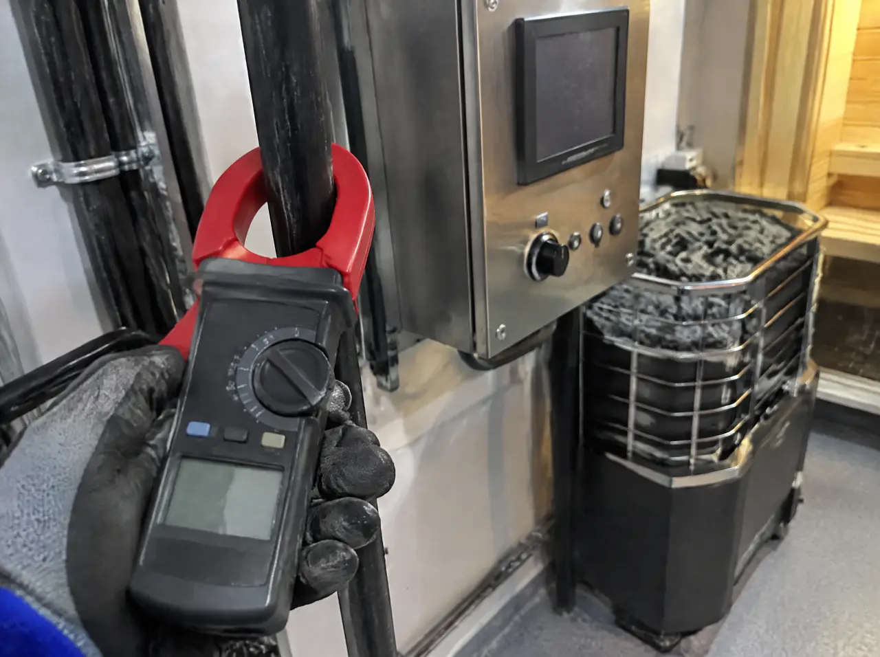 Close-up of an electrician's clamp meter around a thick black cable in a tidy plant room beside a sauna heater controller