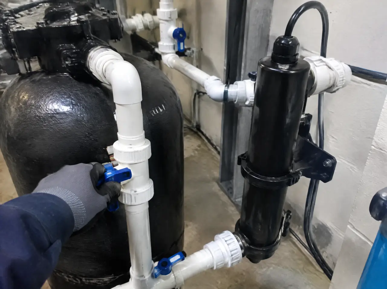 Plant-room close-up of a filtration vessel and a UV unit in plain black casings, white PVC pipework and valves, and a technician's gloved hand on a valve