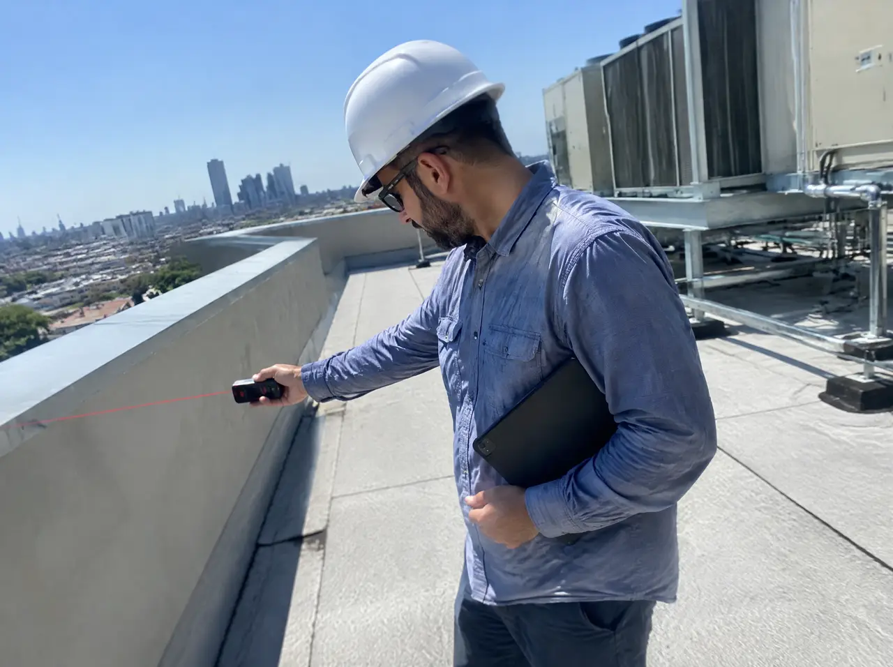 Project engineer in a hard hat measuring a rooftop terrace with a laser measure in harsh midday sun, a tablet under his arm