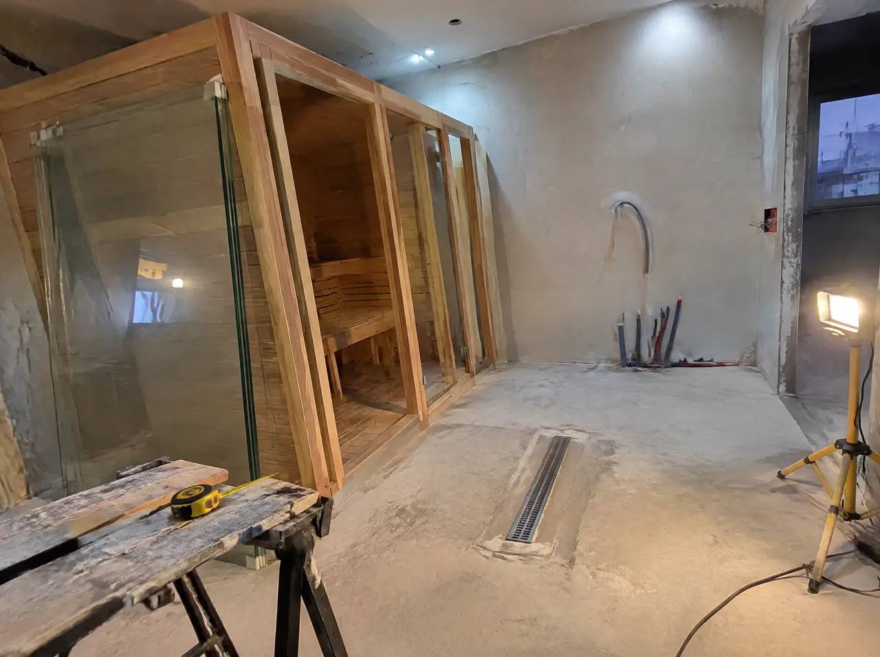 Half-finished villa wellness room in Riyadh with plaster walls, a cedar cabin frame with glass panels leaning against it, conduit and a floor drain, and a tape measure on a trestle